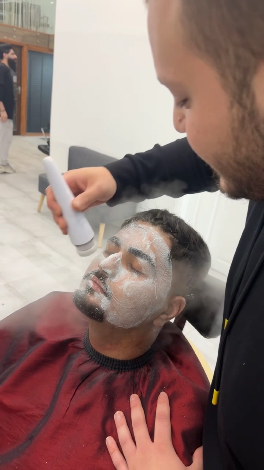 A barber applying a white face mask treatment to a reclined client in a red gown
